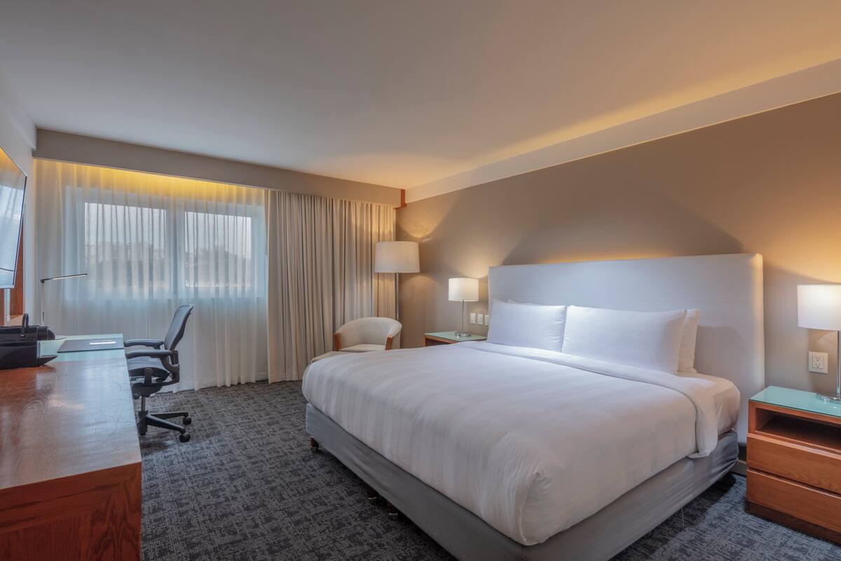 Habitación de hotel con una cama grande, un escritorio con silla, una lámpara y una ventana con cortinas.