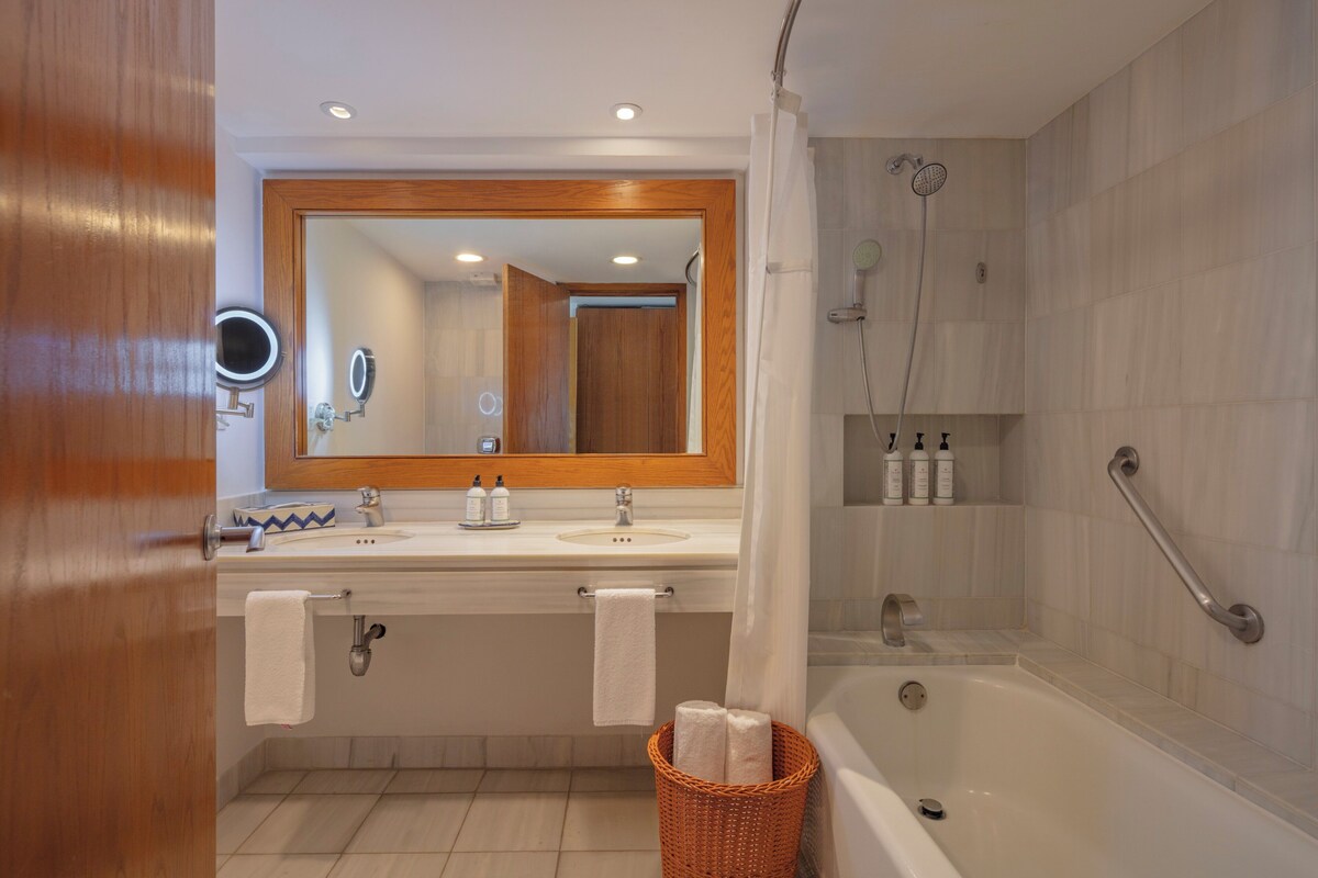 Un baño con puerta de madera, un espejo grande, dos lavamanos, una bañera y una ducha con cabezal de ducha manual.