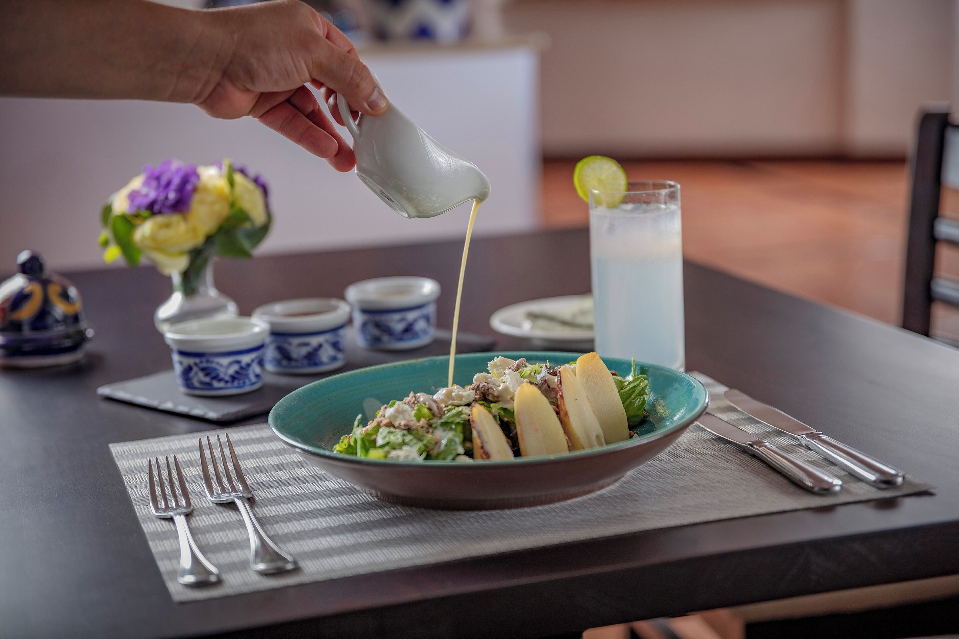 Ein Tisch mit einer Salatschüssel, einem Glas Eistee und einer Hand, die ein Dressing darüber gießt.