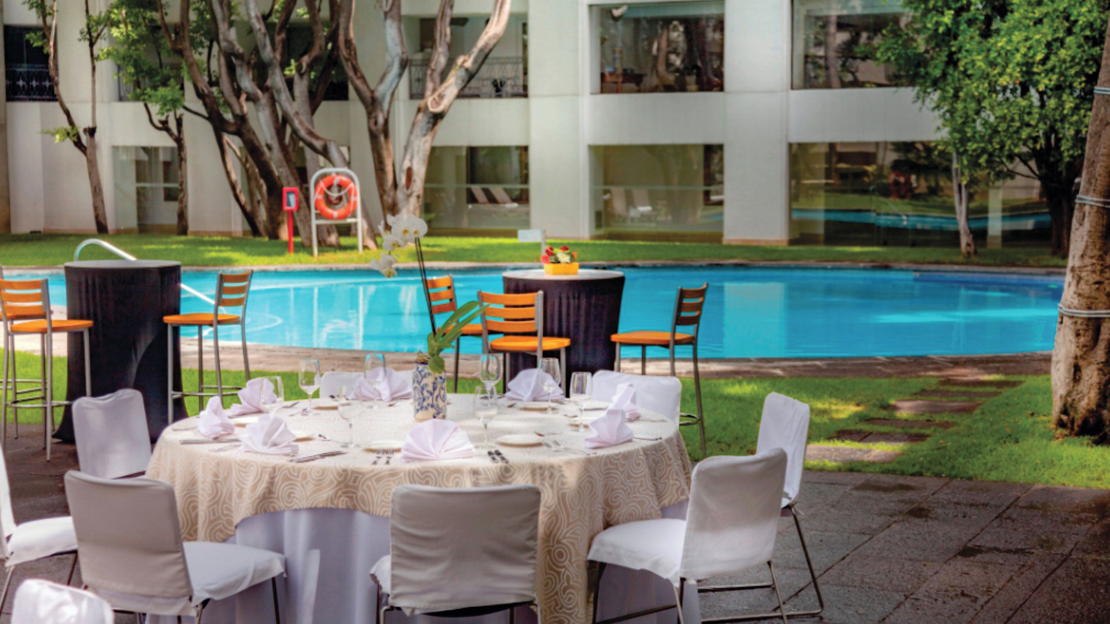 An outdoor dining area with tables set for a meal, chairs, and a pool in the background.