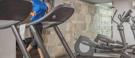 Un gimnasio con cintas de correr y elípticas, una persona usando una cinta y una pared de piedra al fondo.