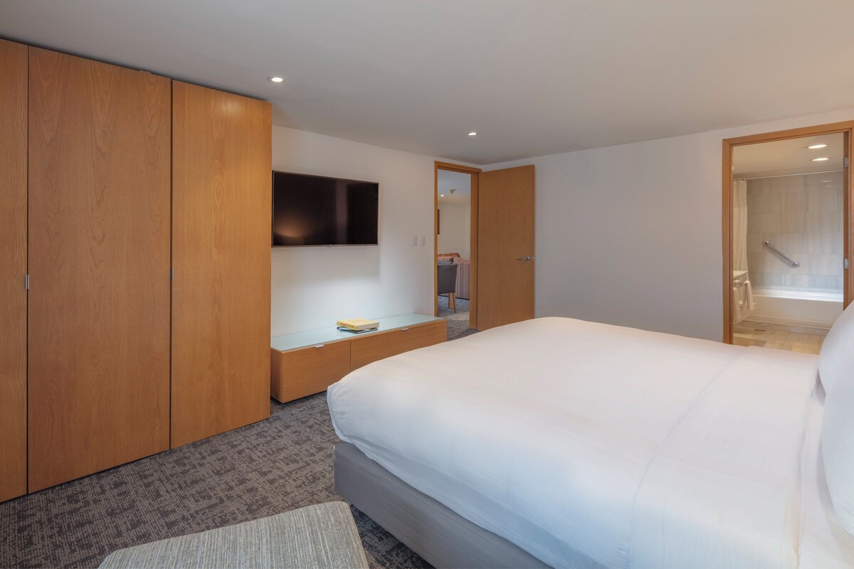 Habitación de hotel con una cama grande, armario de madera, televisor de pantalla plana en la pared y una mesita de noche pequeña.