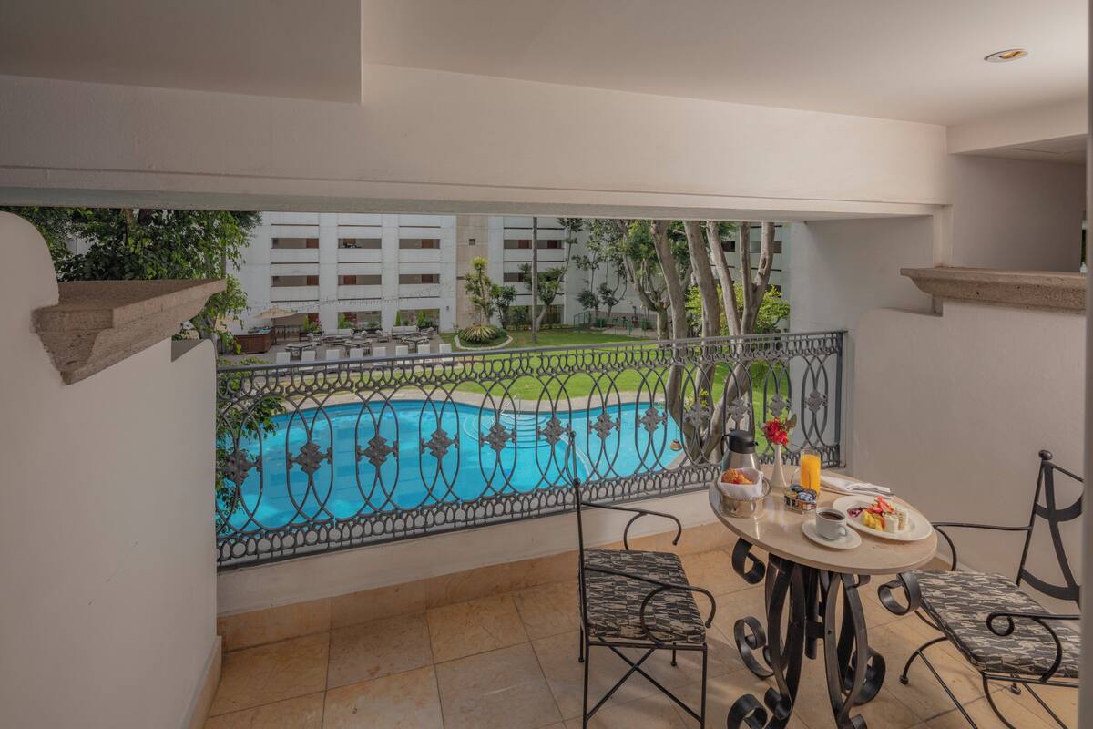 Un balcón con vista a la piscina, mobiliario exterior y una mesa con alimentos para el desayuno.
