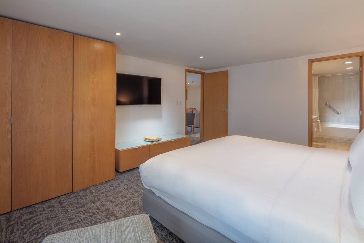 Habitación de hotel con una cama grande, armario de madera, televisor de pantalla plana en la pared y una mesita de noche pequeña.