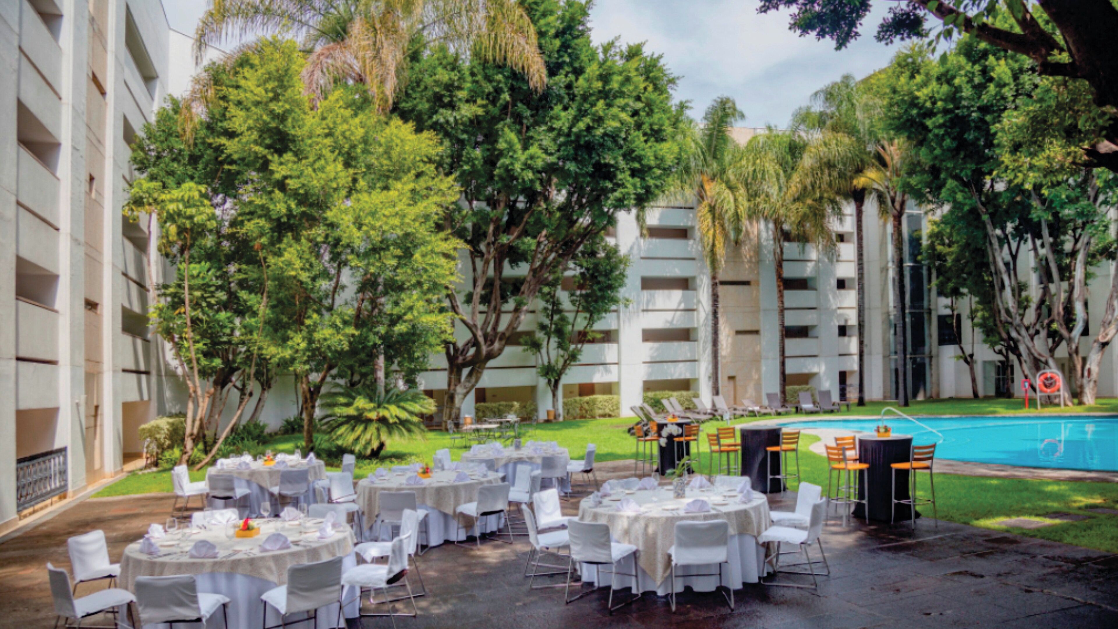 A courtyard with round tables set up for dining, surrounded by trees and a swimming pool.