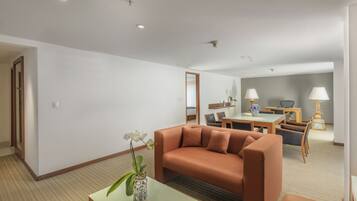 A modern hotel room with a sofa, a small table with a vase of flowers, a dining area with a table and chairs, and a lamp.
