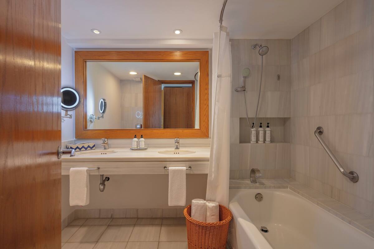 Un baño con puerta de madera, un espejo grande, dos lavamanos, una bañera y una ducha con cabezal de ducha manual.