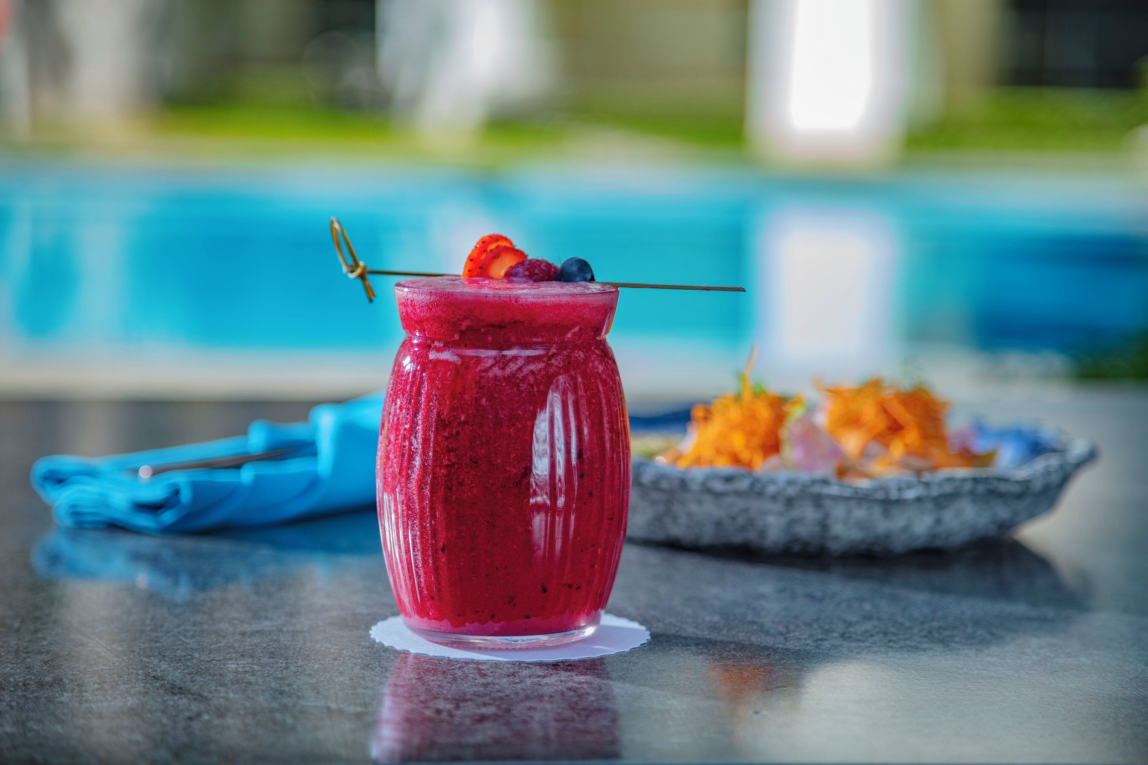 A glass of red smoothie garnished with fruit on a table near a pool.