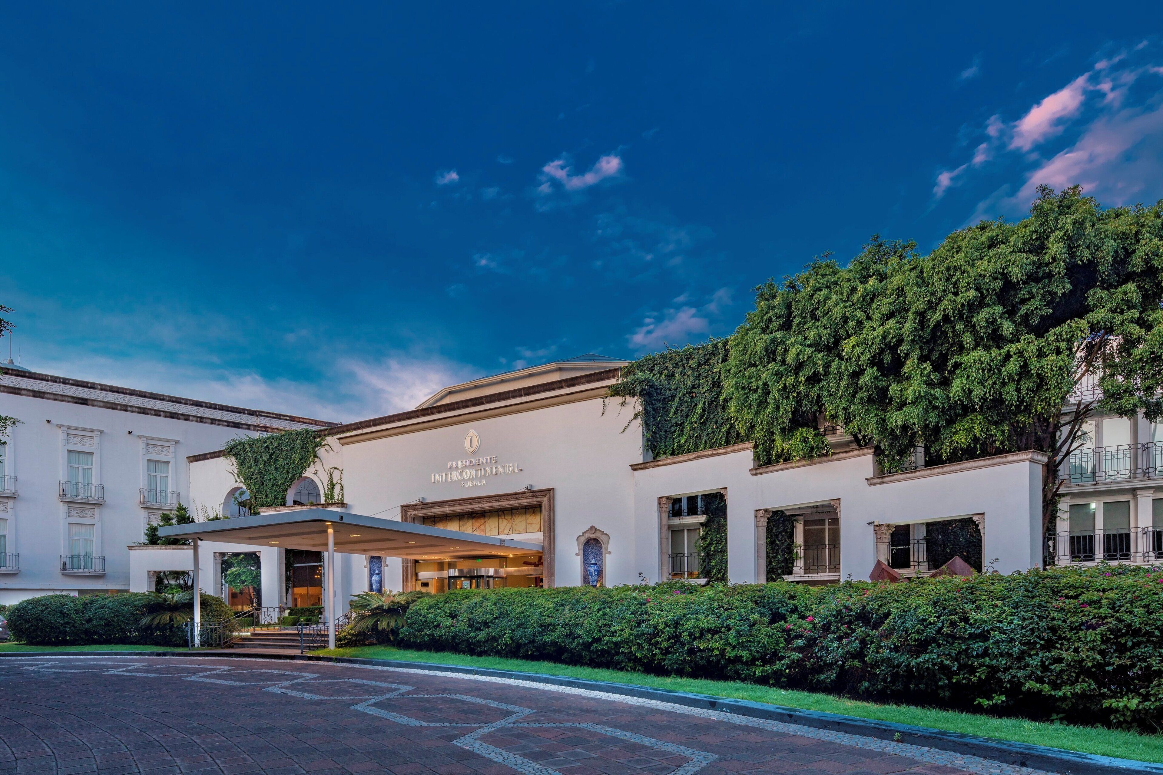 A hotel with a covered entrance, a sign reading 'Presidente Intercontinental Puebla,' and a well-maintained garden.