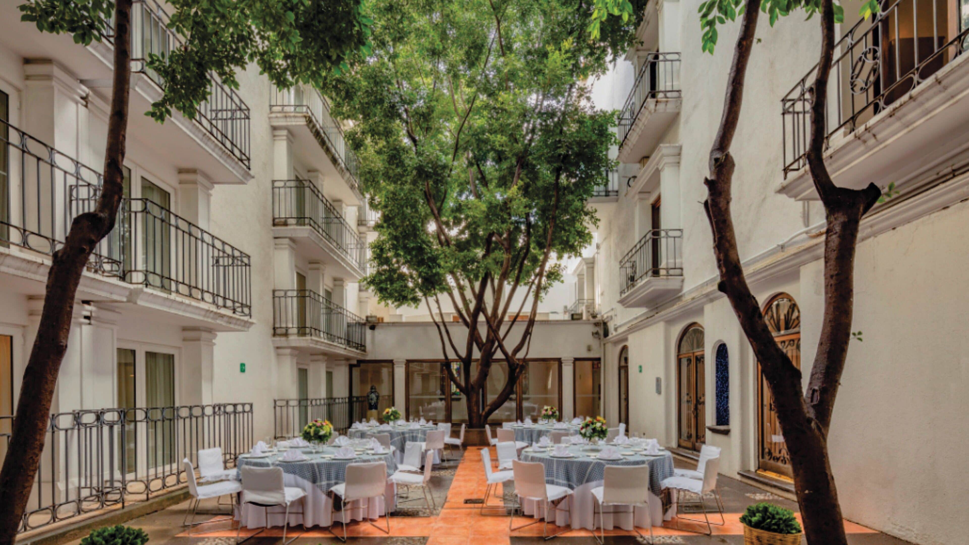 A courtyard with a central dining area surrounded by white buildings, featuring balconies and a tree providing shade.