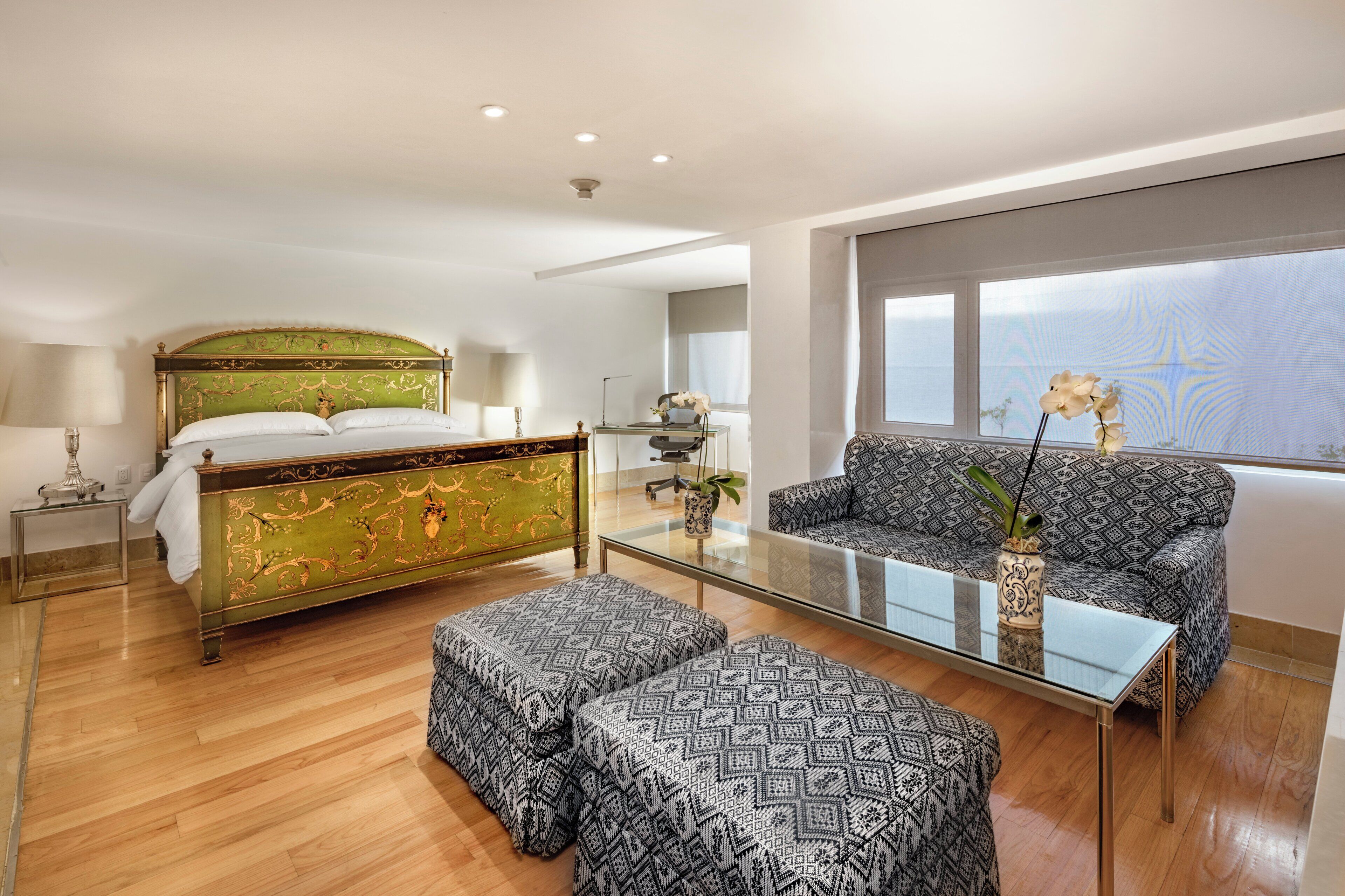 A hotel room with a bed, a sofa, a glass coffee table, and a view of the sky.