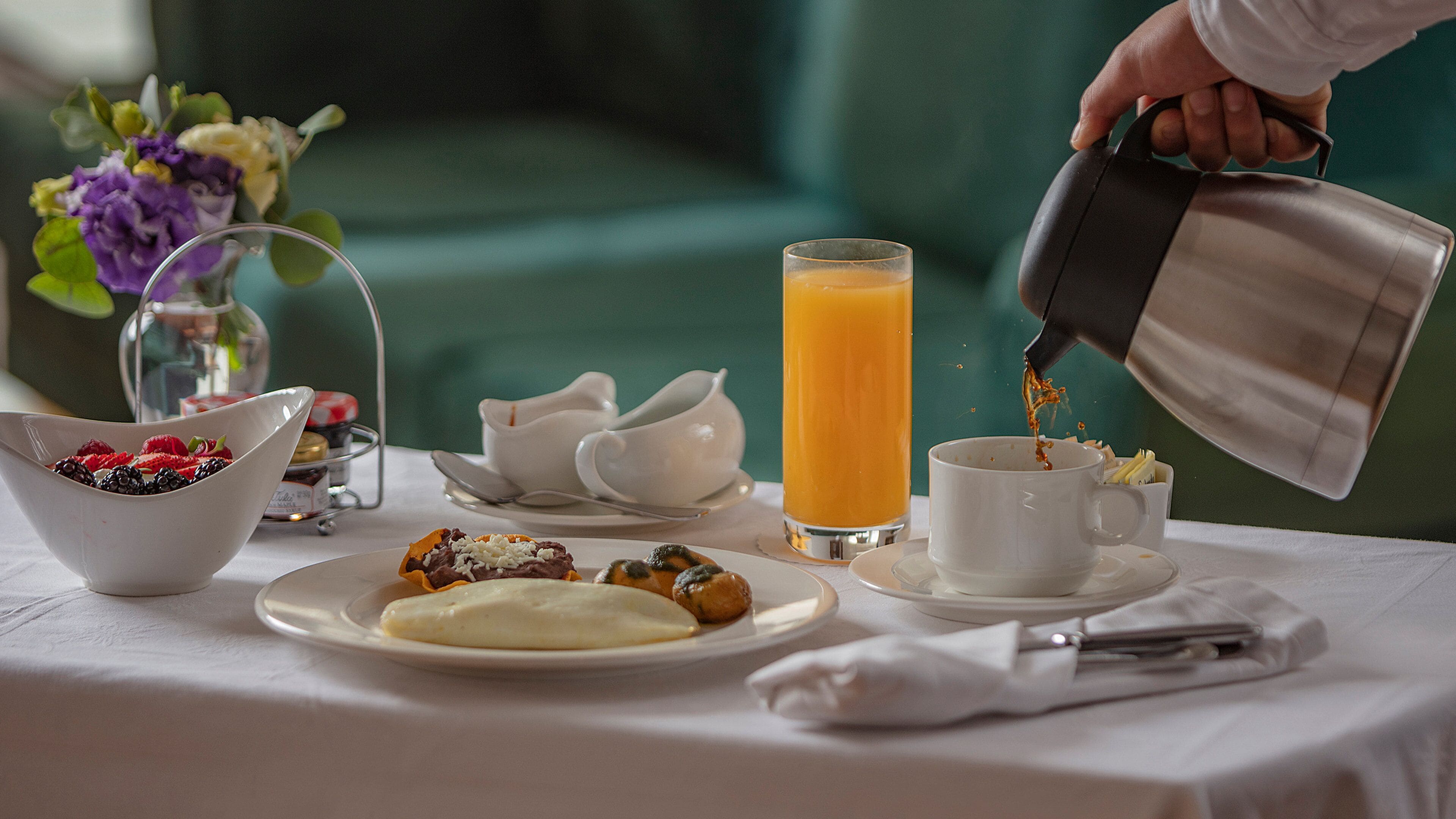 A breakfast setting with a bowl of fruit, a glass of orange juice, and a cup of coffee being poured.