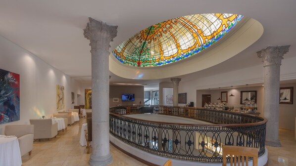 Un vestíbulo espacioso con techo alto, columnas ornamentales, una cúpula de vidrio central y una zona de bar con asientos.