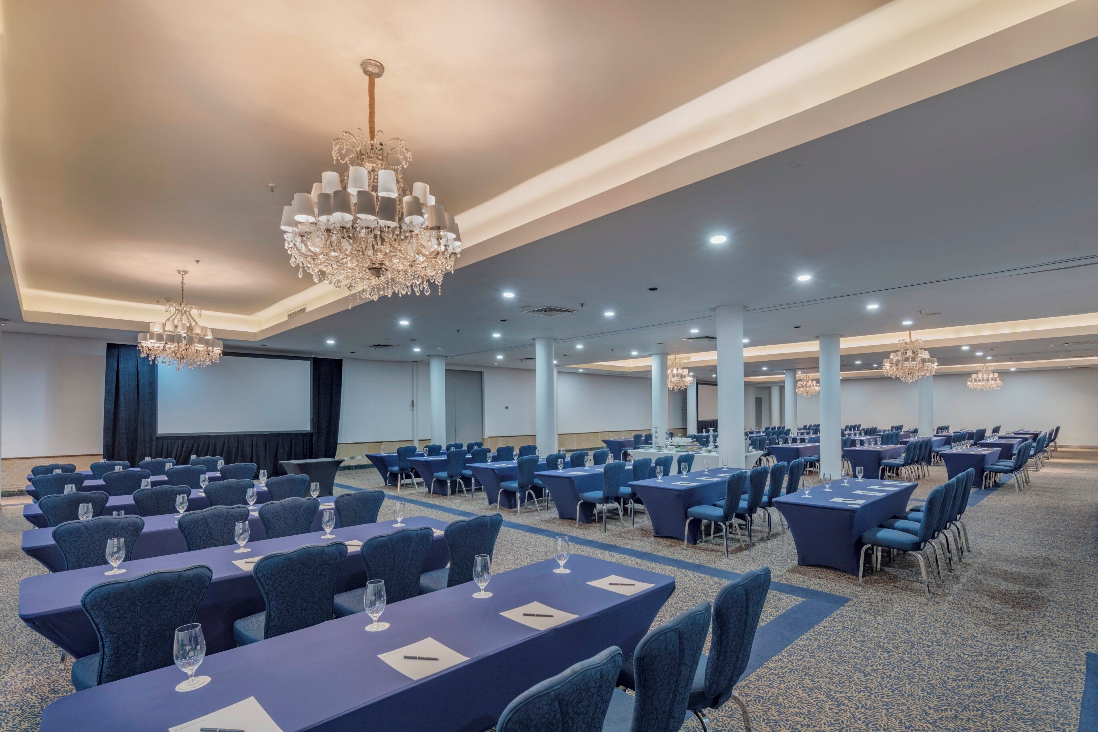 A conference room with multiple tables and chairs, chandeliers, and a large screen.