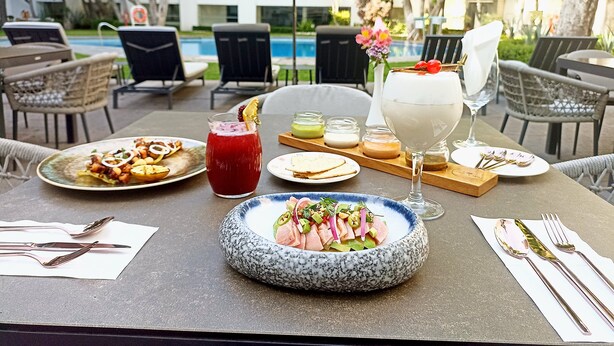 Un área de comedor al aire libre con vista a la piscina, con una mesa dispuesta con diversos platos, bebidas y cubiertos.