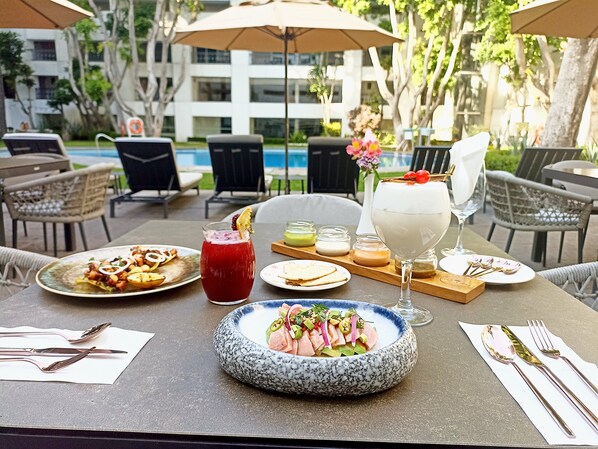 Un área de comedor al aire libre con vista a la piscina, con una mesa dispuesta con diversos platos, bebidas y cubiertos.