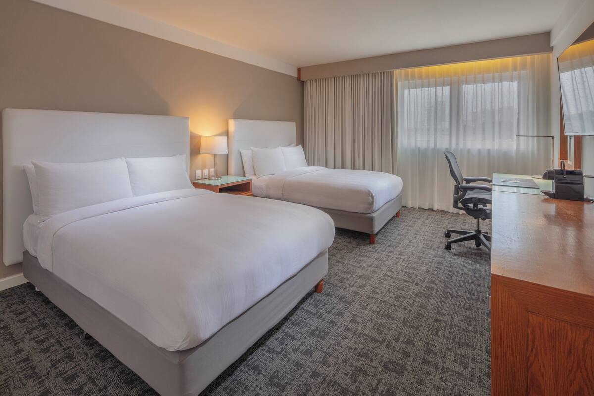 Habitación de hotel con dos camas, un escritorio, una silla y un ventanal con cortinas.