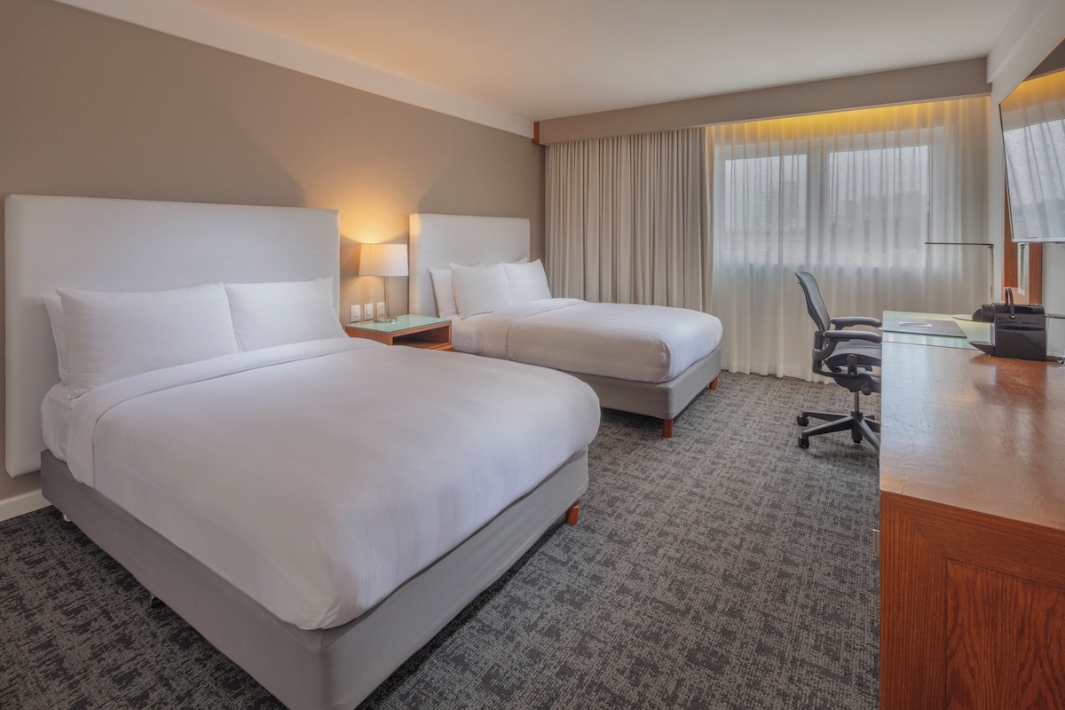 Habitación de hotel con dos camas, un escritorio, una silla y un ventanal con cortinas.