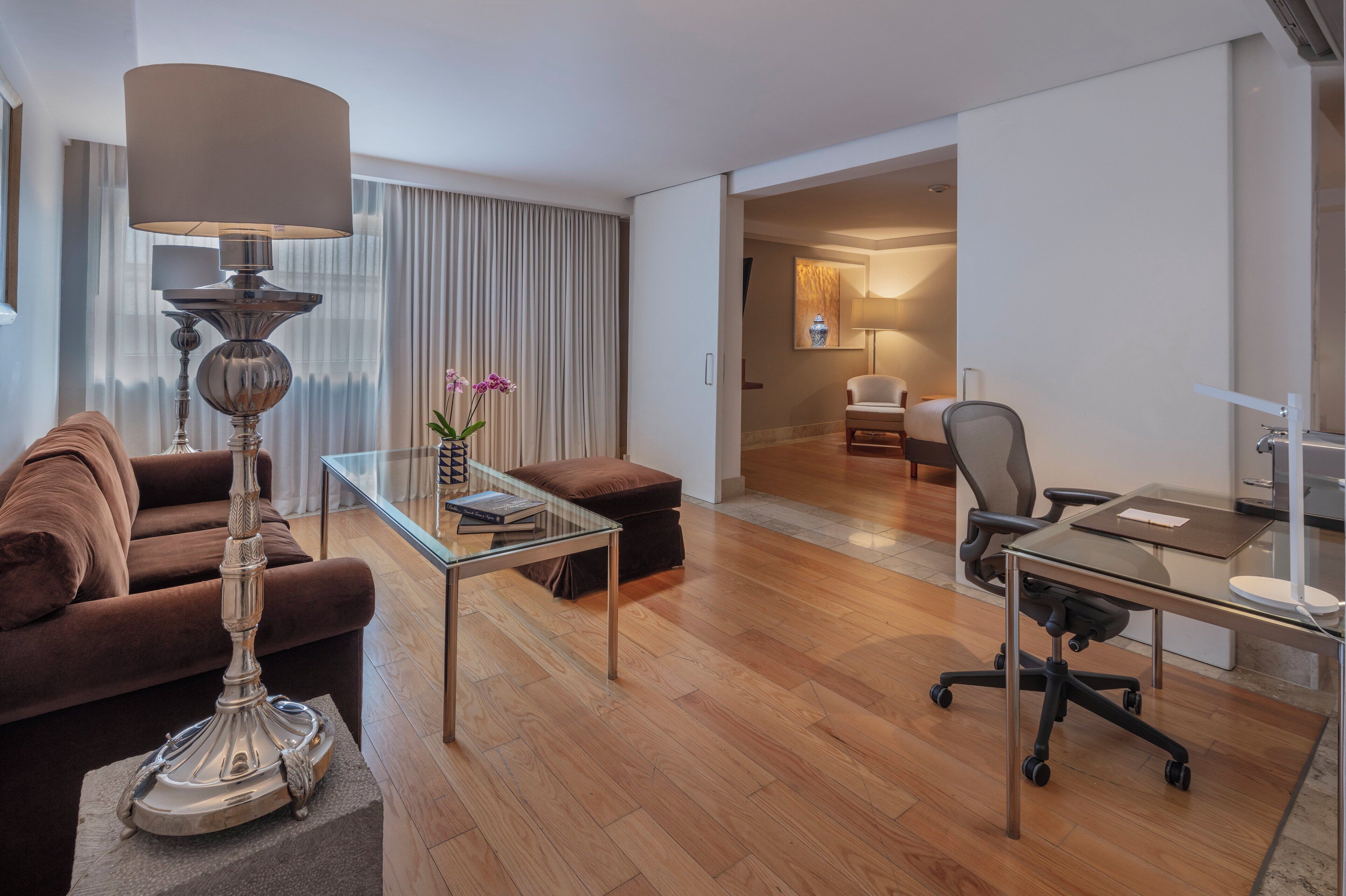 A modern hotel room with a brown sofa, a glass coffee table, a desk with a chair, and a lamp.