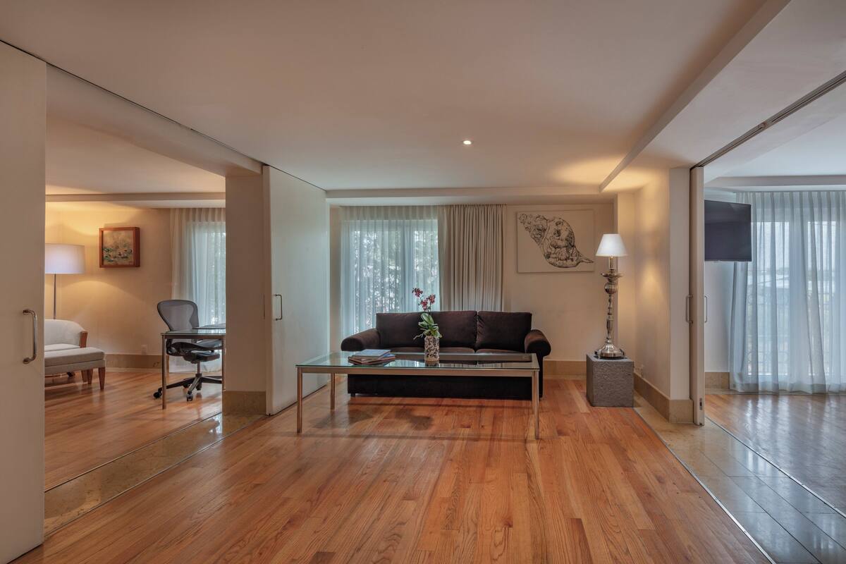 Una habitación de hotel moderna con suelo de madera, un sofá, una mesa de centro con un jarrón, un escritorio con una silla y un televisor.