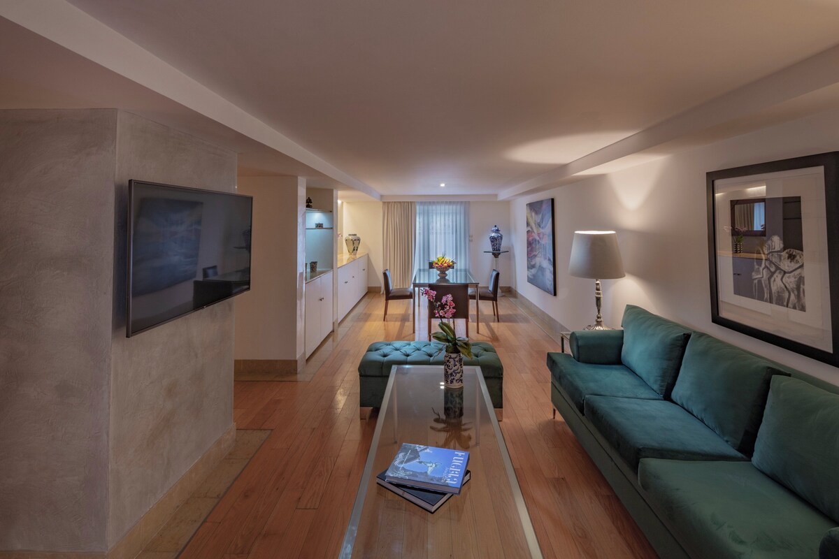 Una sala de estar moderna con un sofá verde, una mesa de centro de vidrio y una zona de comedor al fondo.