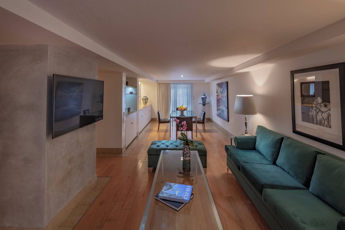 Una sala de estar moderna con un sofá verde, una mesa de centro de vidrio y una zona de comedor al fondo.