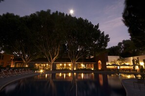 Un espace aménagé au bord de la piscine, avec des chaises longues, et un bâtiment dont les fenêtres sont illuminées au crépuscule.
