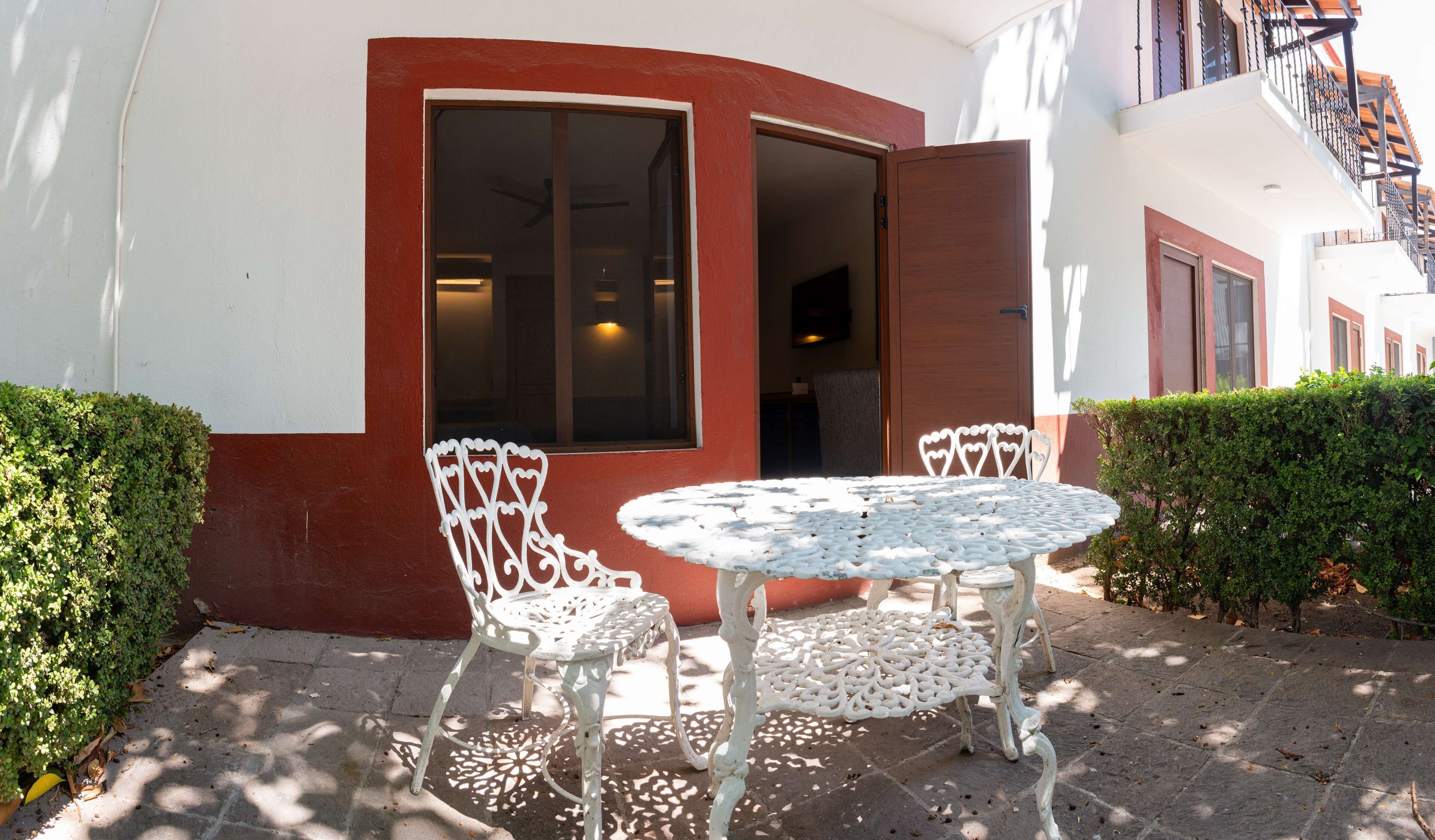 A white outdoor seating area with a table and two chairs, a red and white building with a large window and door, and a staircase visible in the background.