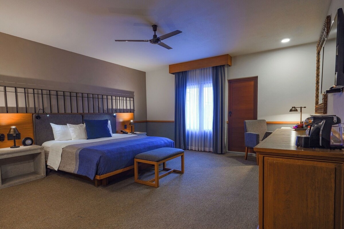Habitación de hotel con cama, escritorio, silla, cafetera, televisor y ventilador de techo.