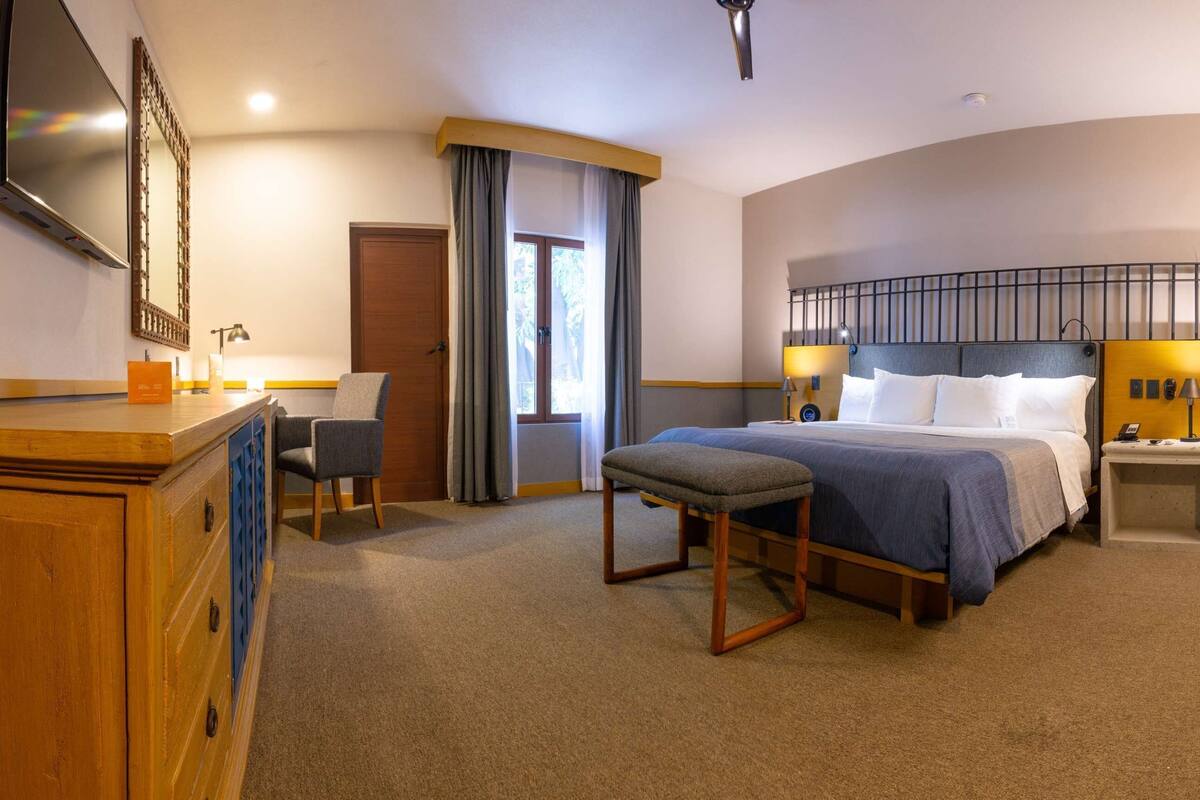 Habitación de hotel con cama, mesitas de noche, escritorio, silla, televisor y un gran ventanal con cortinas.