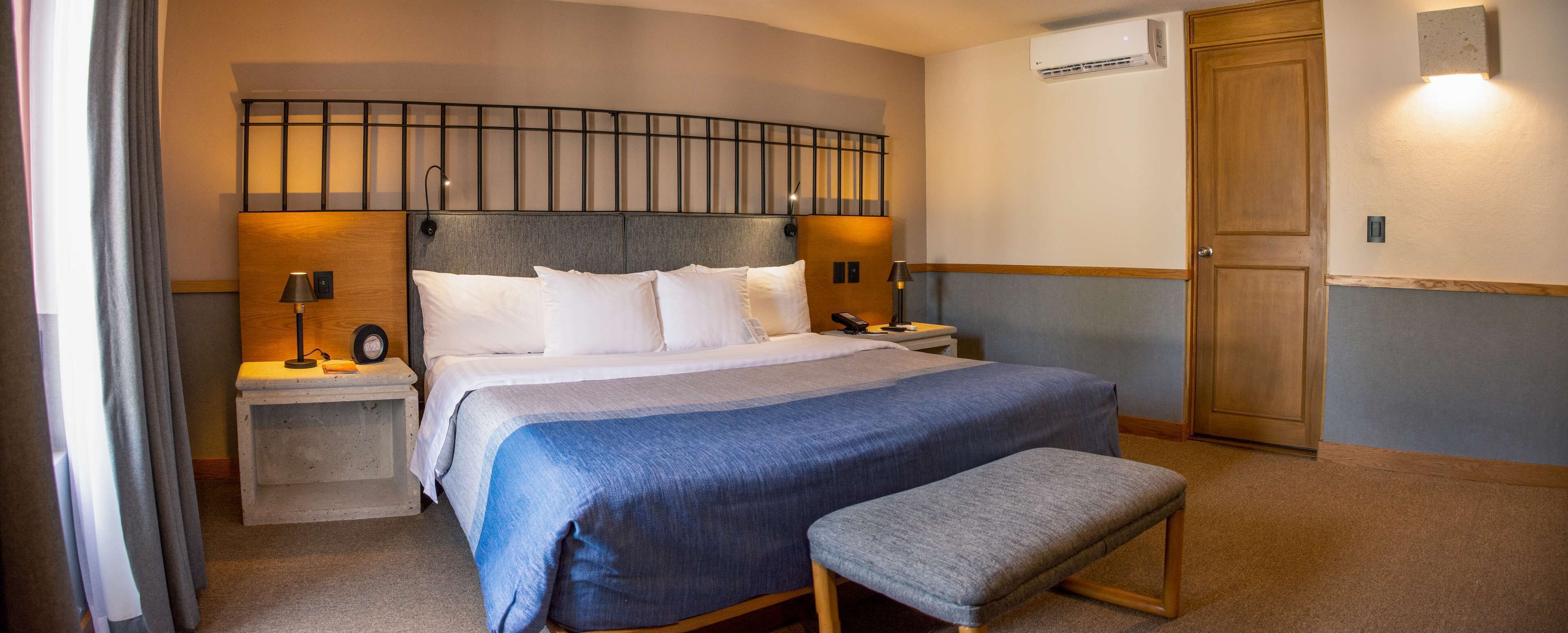 Una habitación de hotel ordenada con una cama grande, mesitas de noche, un banco, un equipo de aire acondicionado y una puerta de madera.