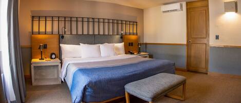 Una habitación de hotel ordenada con una cama grande, mesitas de noche, un banco, un equipo de aire acondicionado y una puerta de madera.