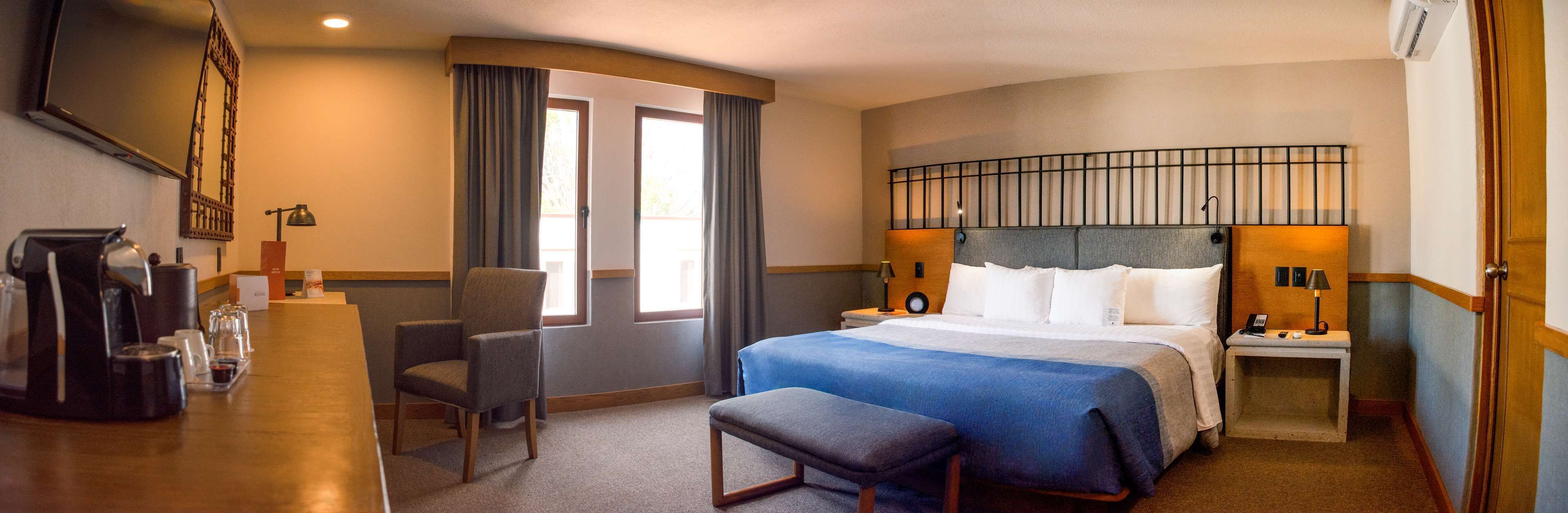 A hotel room with a bed, a desk with a coffee maker, a chair, and a window with curtains.