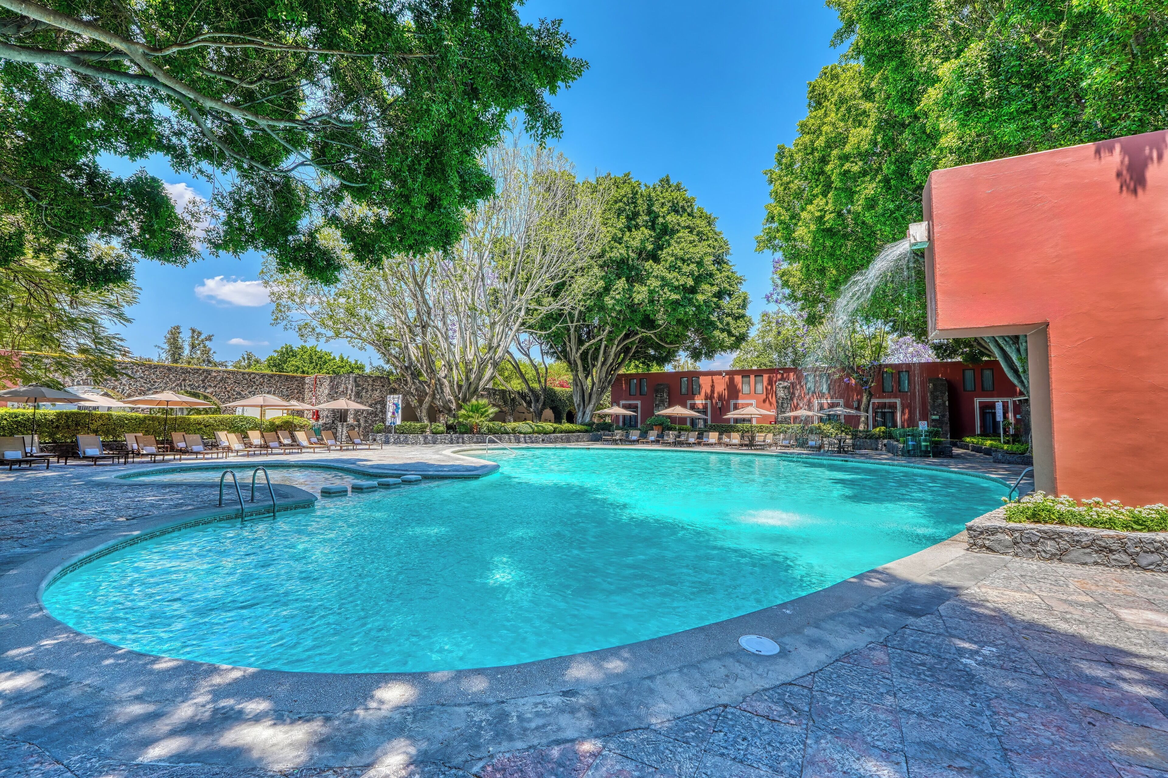 A swimming pool with lounge chairs and umbrellas, surrounded by trees and a building.