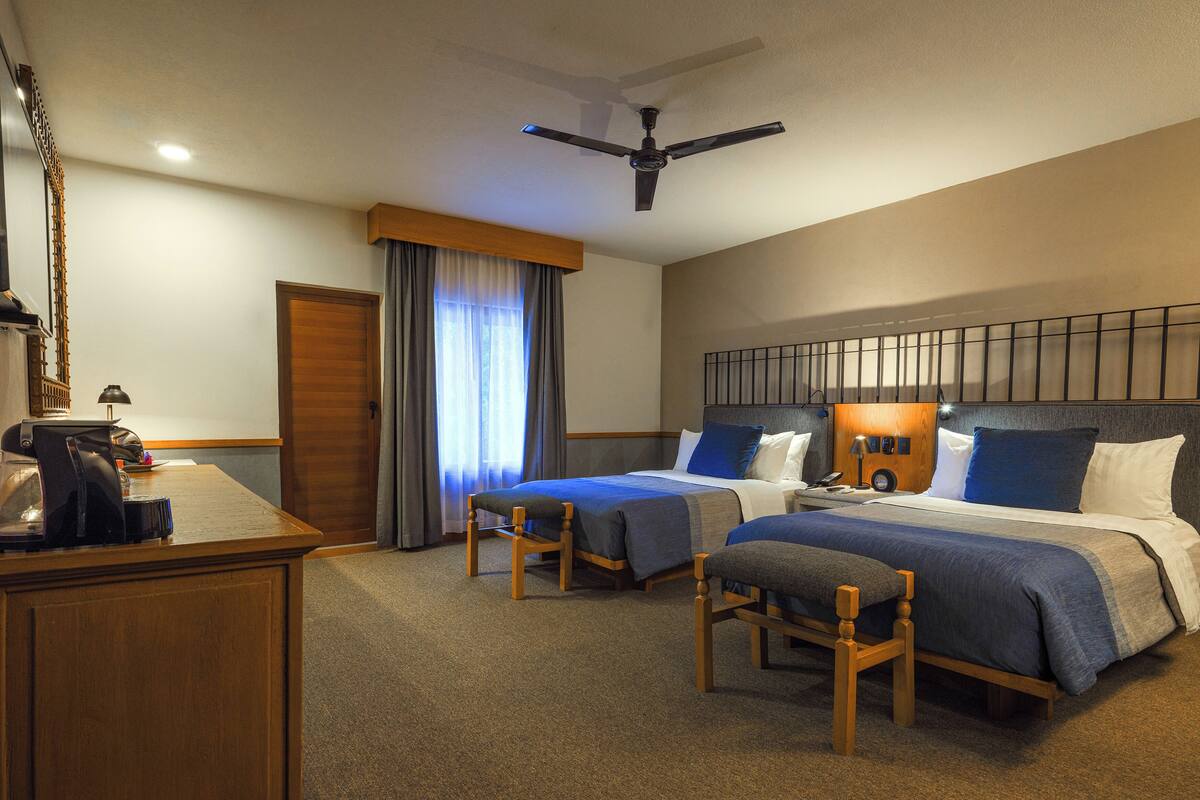 Habitación de hotel con dos camas, un escritorio, una silla, un ventilador de techo y una ventana con cortinas.