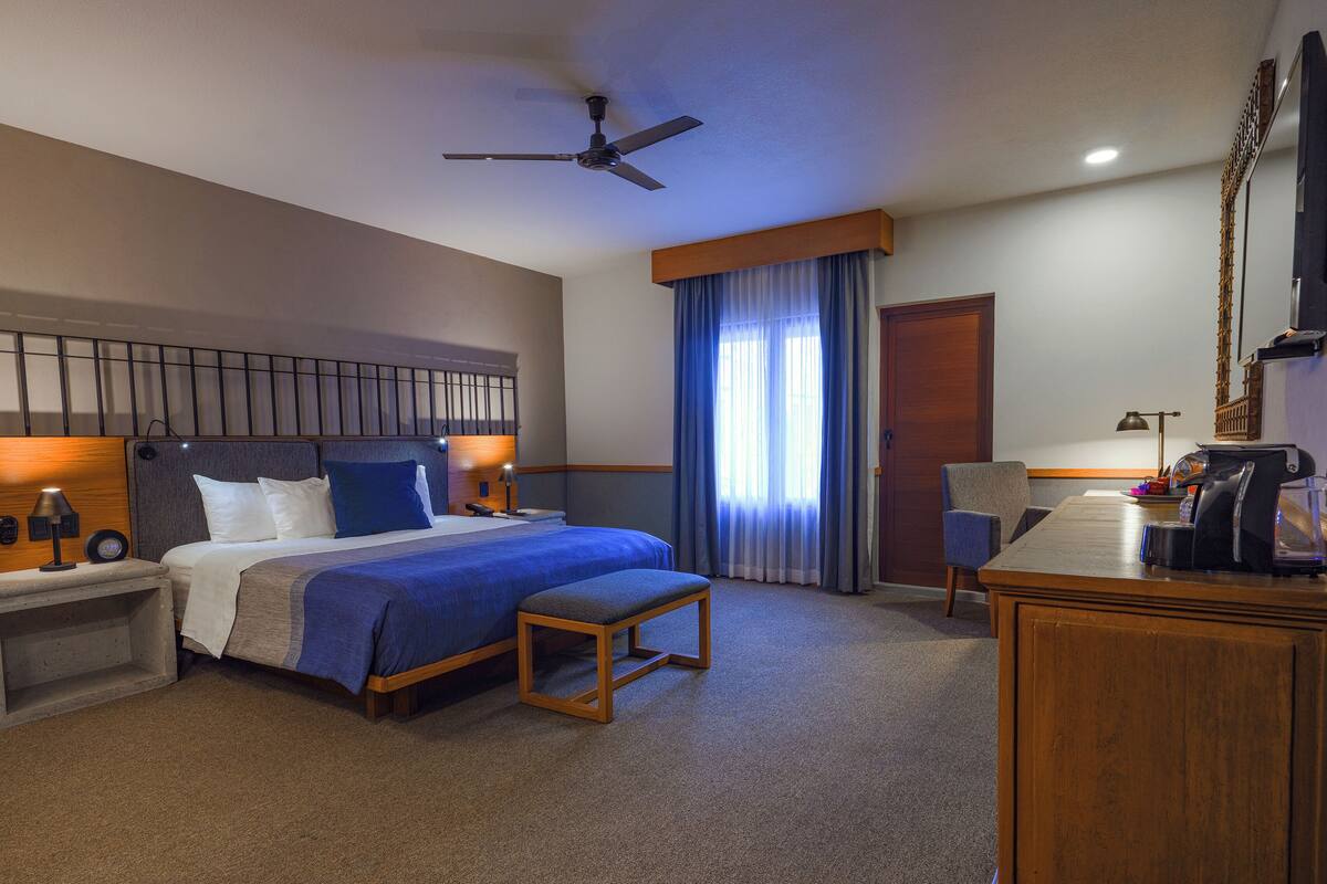 Habitación de hotel con cama, mesita de noche, lámpara, silla, escritorio y ventilador de techo.