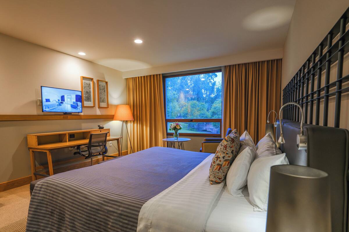 Habitación de hotel con cama, escritorio, silla, televisor y ventana con cortinas.