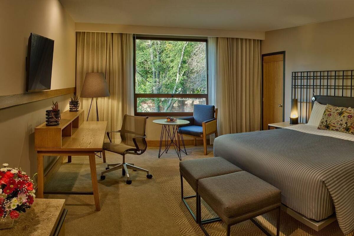Una habitación de hotel con cama, mesitas de noche, escritorio, silla, una pequeña mesa redonda y ventana con cortinas.