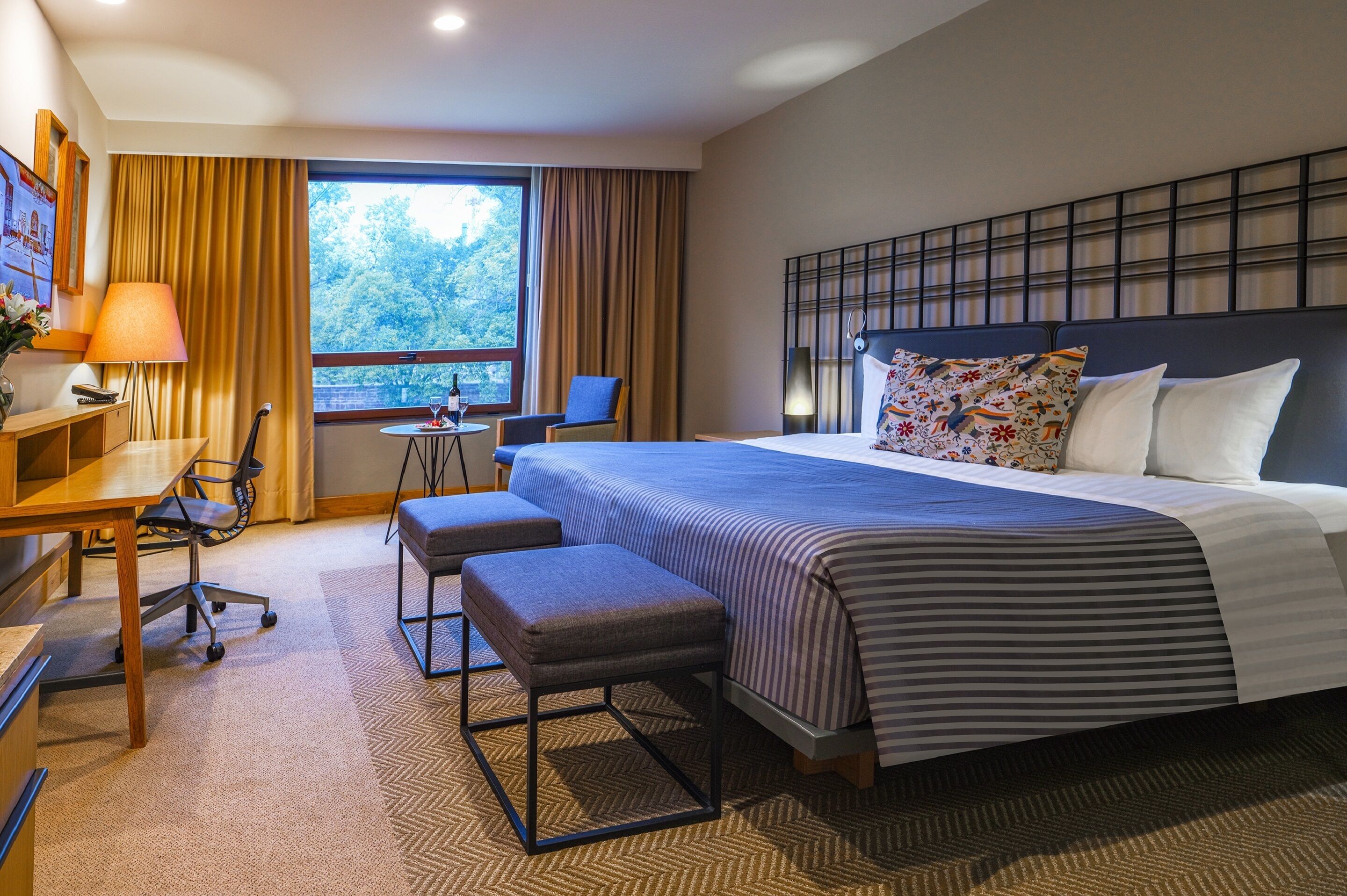 A hotel room with a large bed, a desk with a chair, a small table with a chair, and a window with curtains.