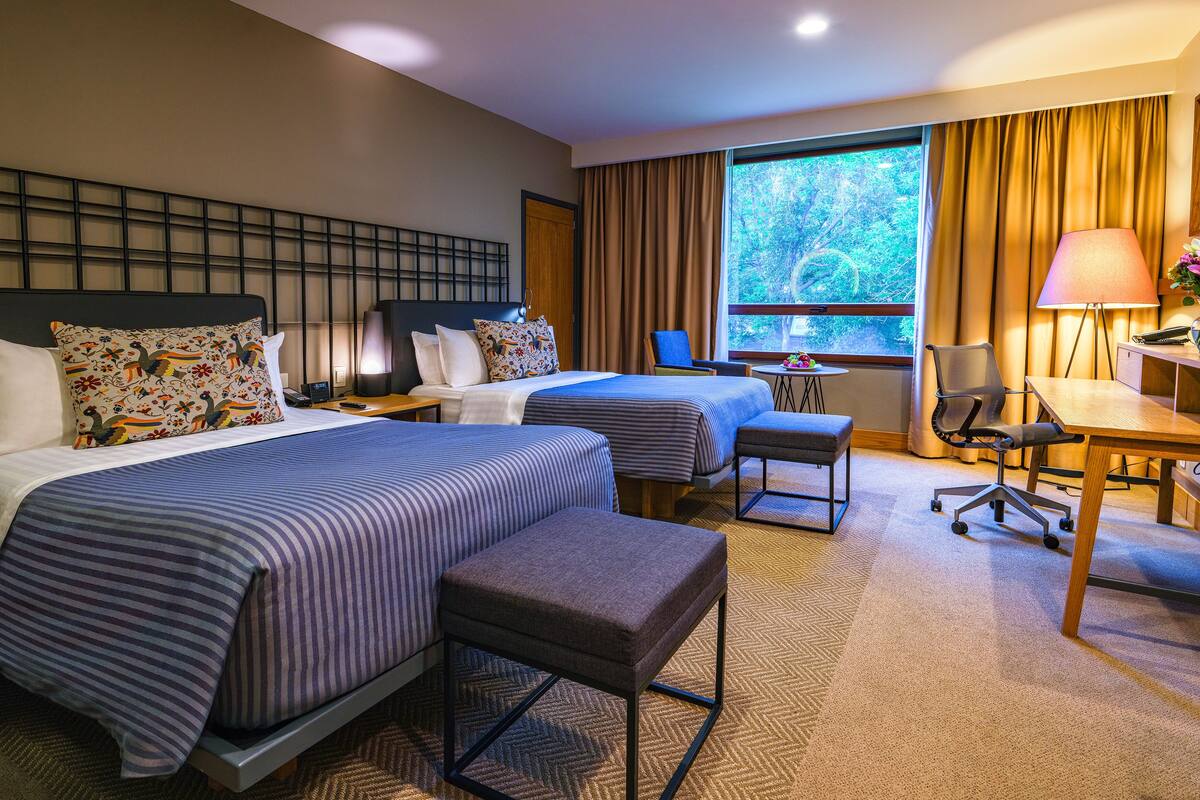 Habitación de hotel con dos camas, un escritorio, una silla, una lámpara y vistas a árboles por la ventana.