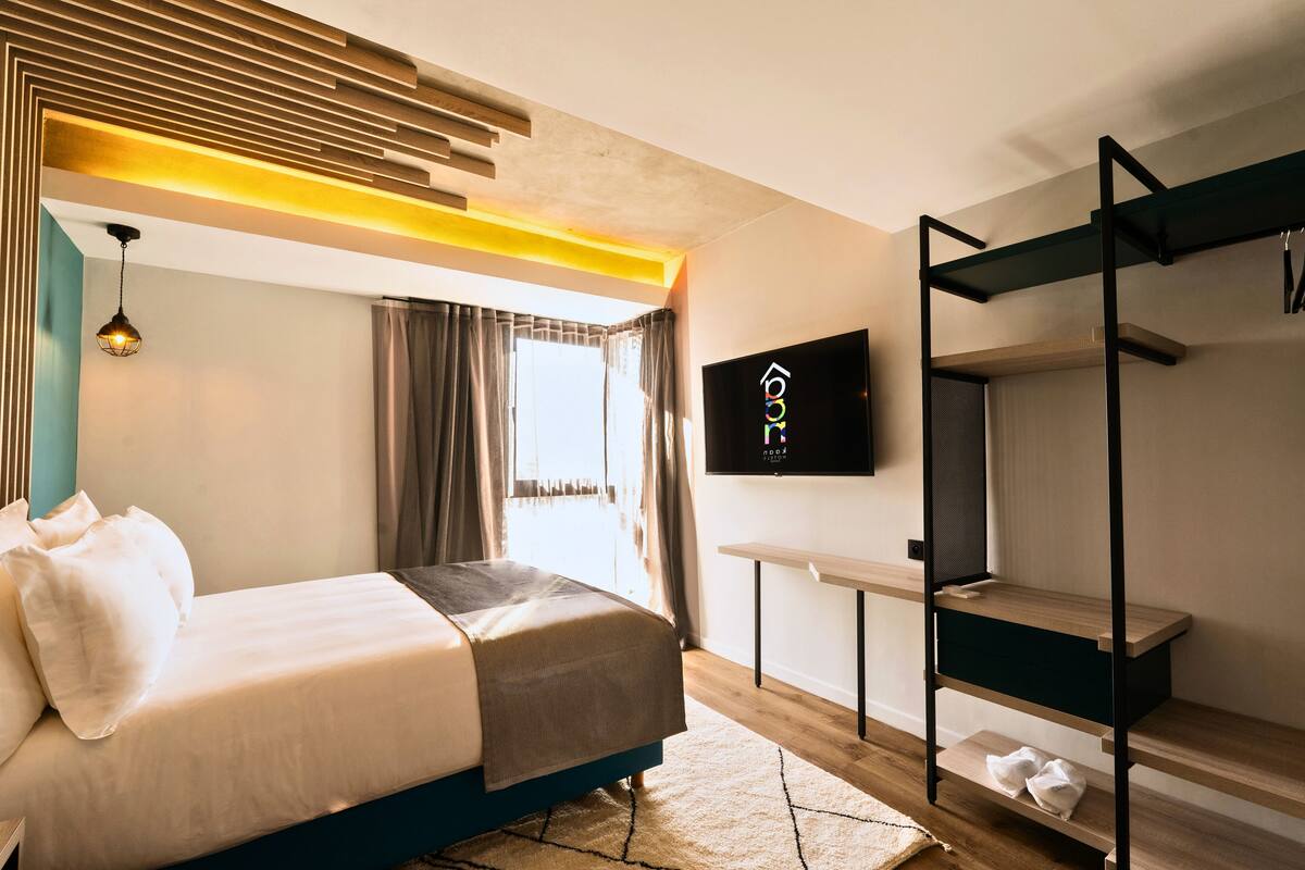 Een moderne hotelkamer met een bed, een aan de muur bevestigde televisie, een houten bankje en een raam met gordijnen.