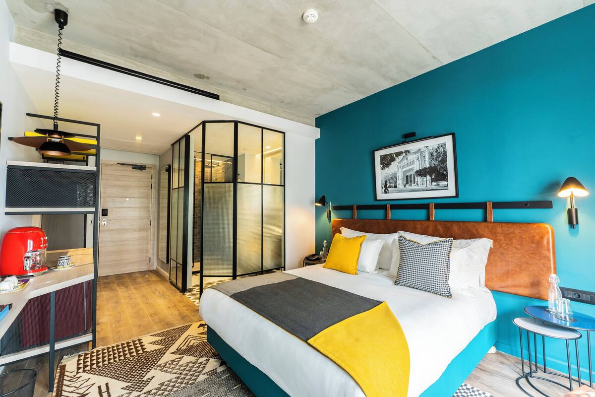 Een moderne hotelkamer met een groot bed, een bureau met koffiezetapparaat, een glazen scheiding en een muur in teal.