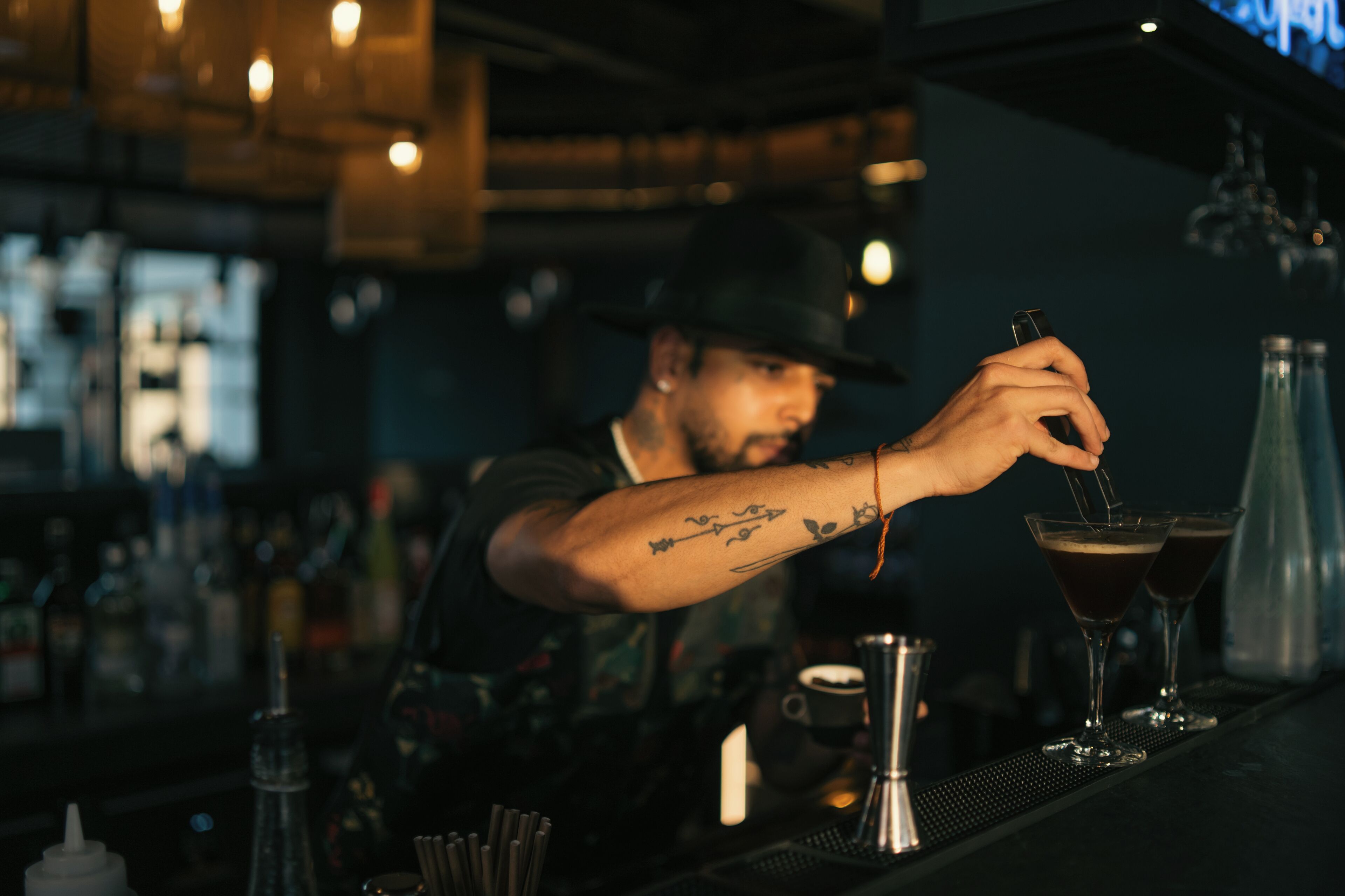 Un barman prépare des boissons dans un bar où l’on peut voir des bouteilles et des verres.