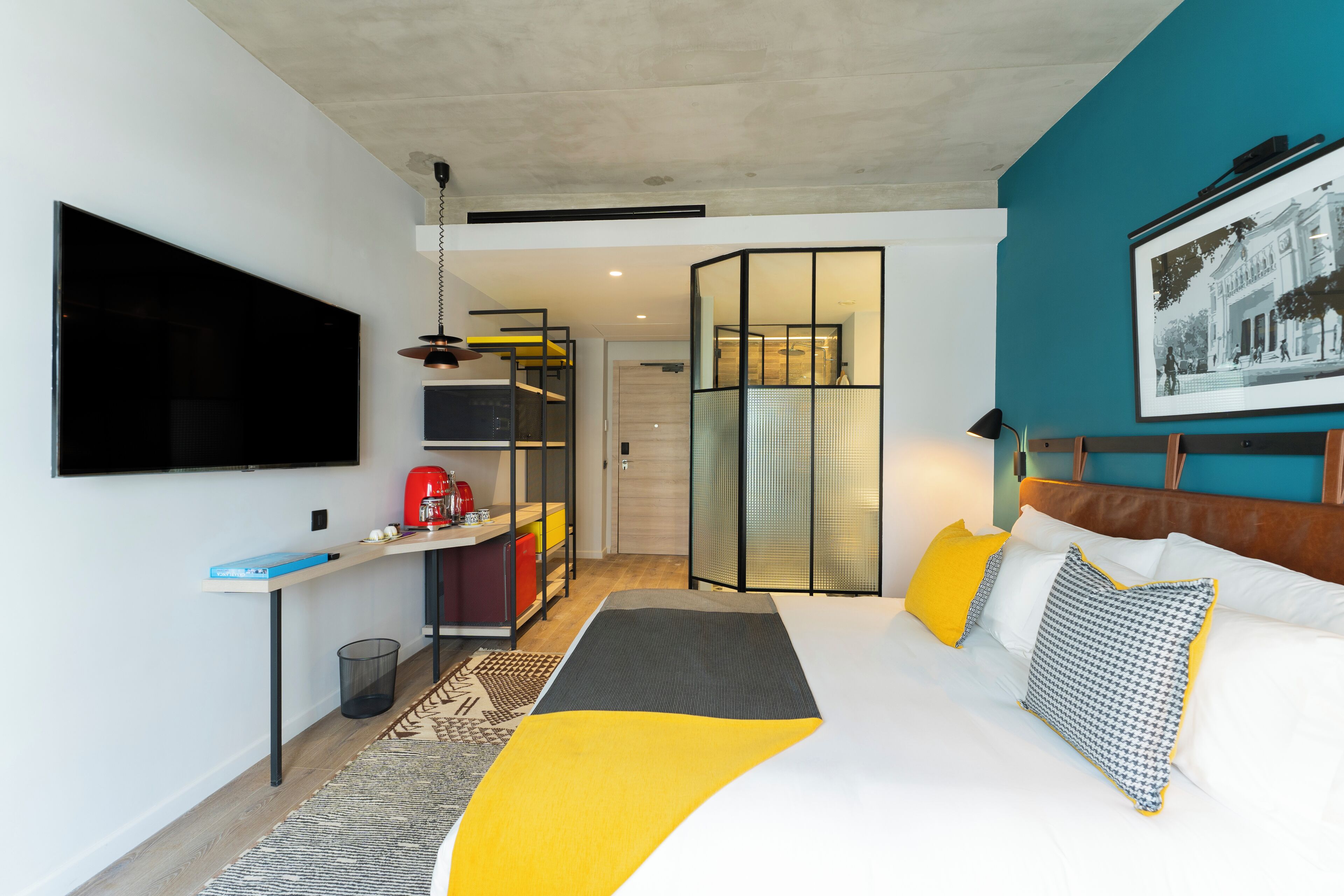 Quarto de hotel moderno com uma cama grande, uma escrivaninha com computador, uma televisão e uma porta de correr para um banheiro.