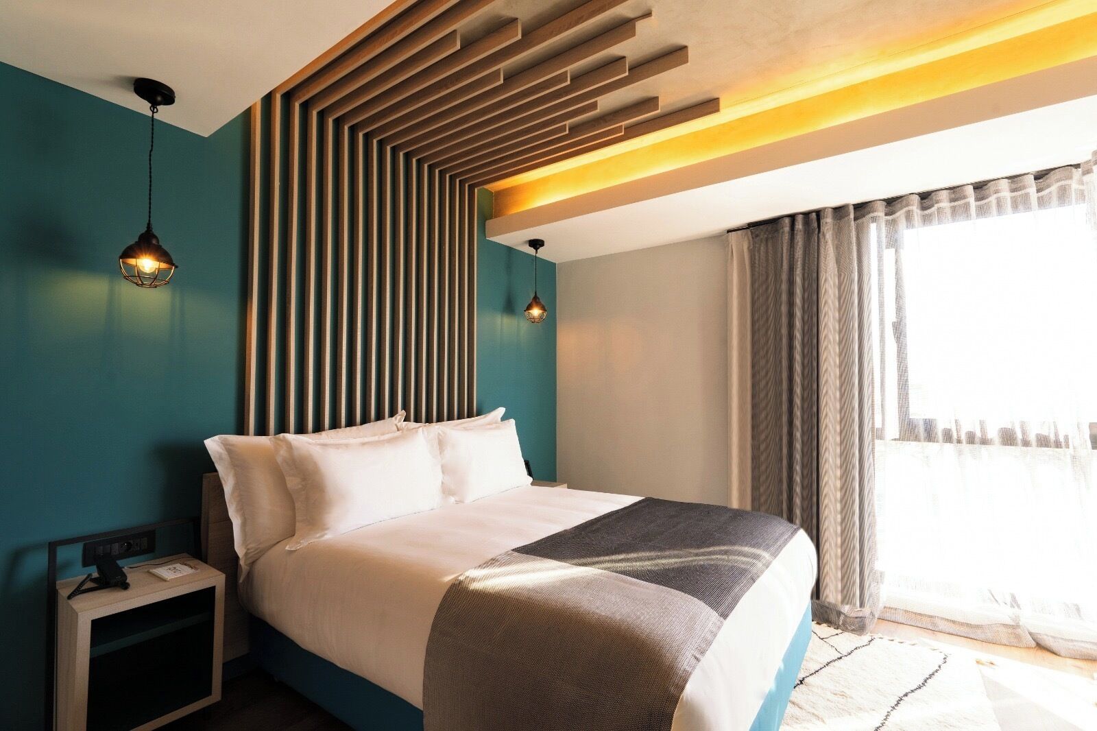 A modern hotel room with a large bed, a nightstand, a wall-mounted light fixture, and a window with curtains.