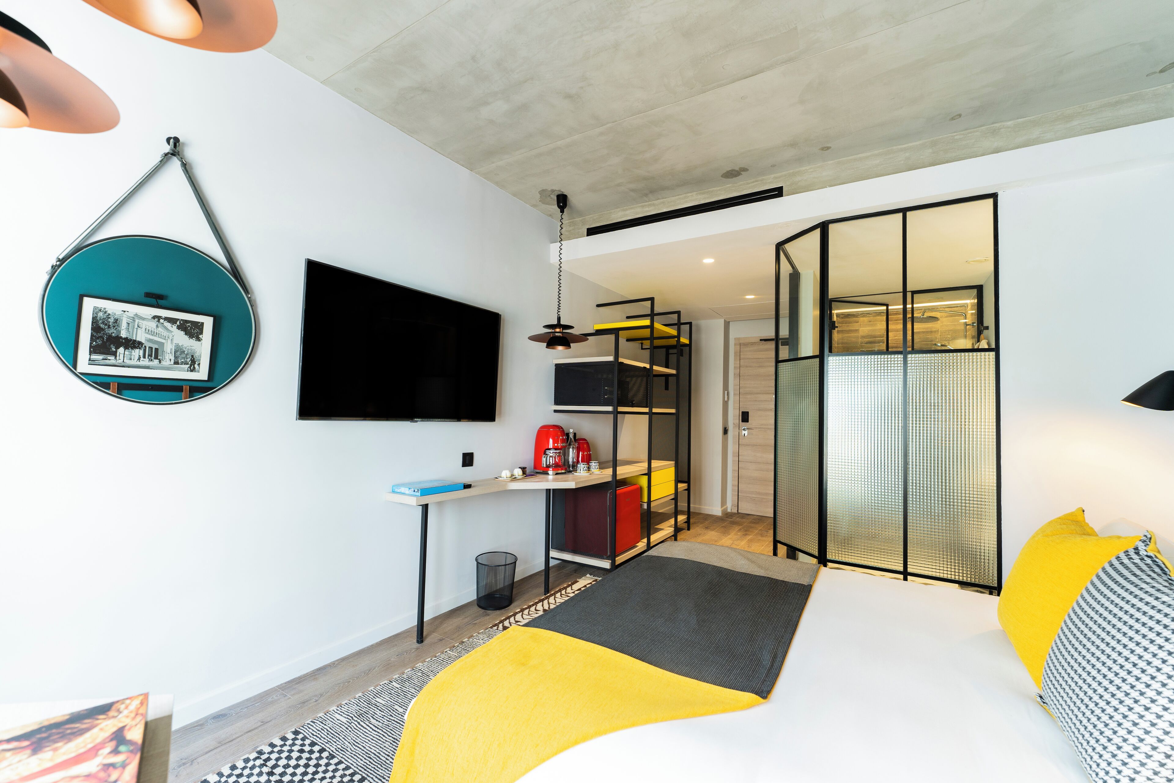 A modern bedroom with a bed, a desk, a TV, and a bathroom visible through a glass partition.