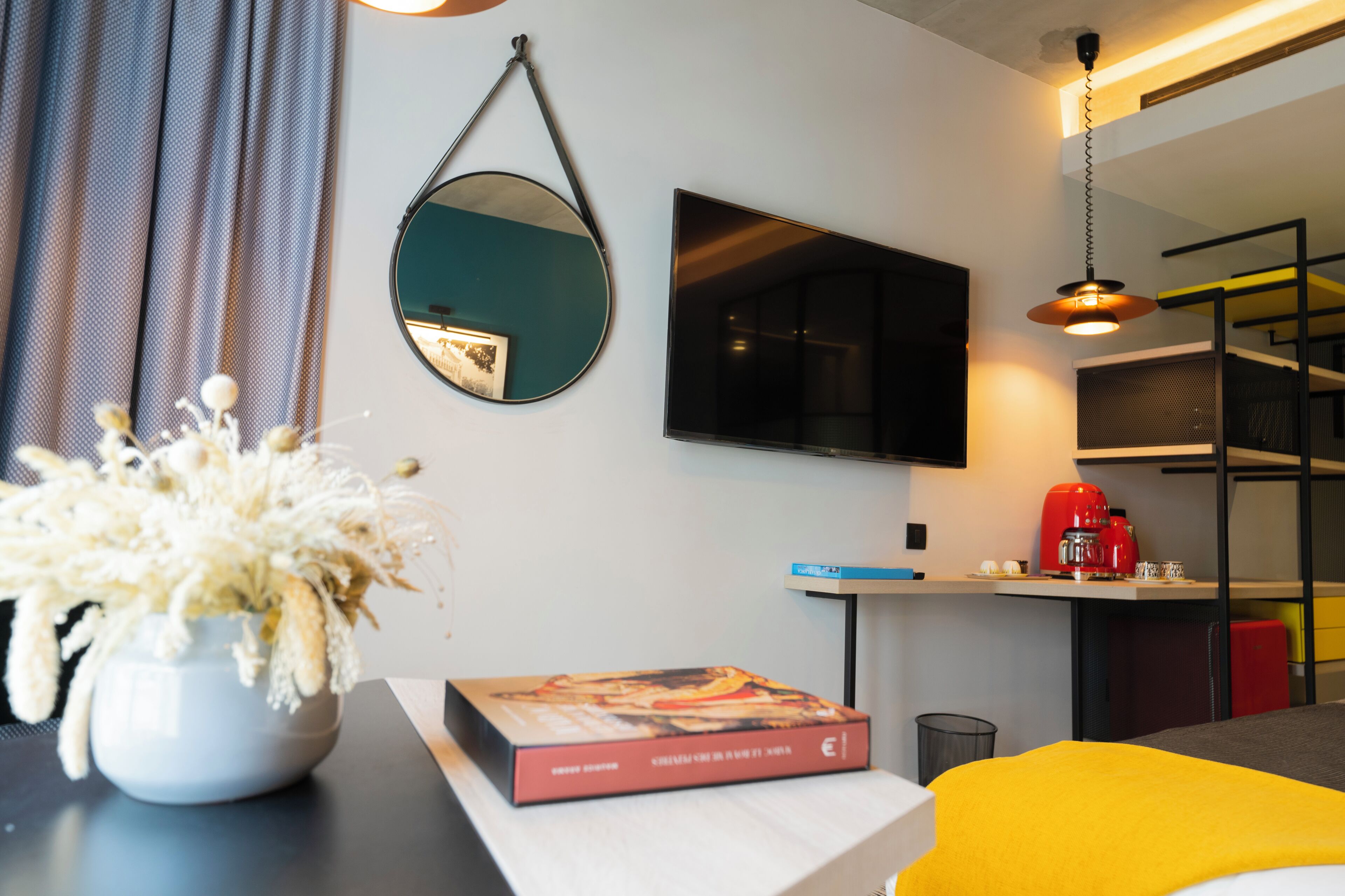 A modern hotel room with a flat-screen TV, a round mirror, a potted plant, a book, a red coffee machine, and a yellow bedspread.