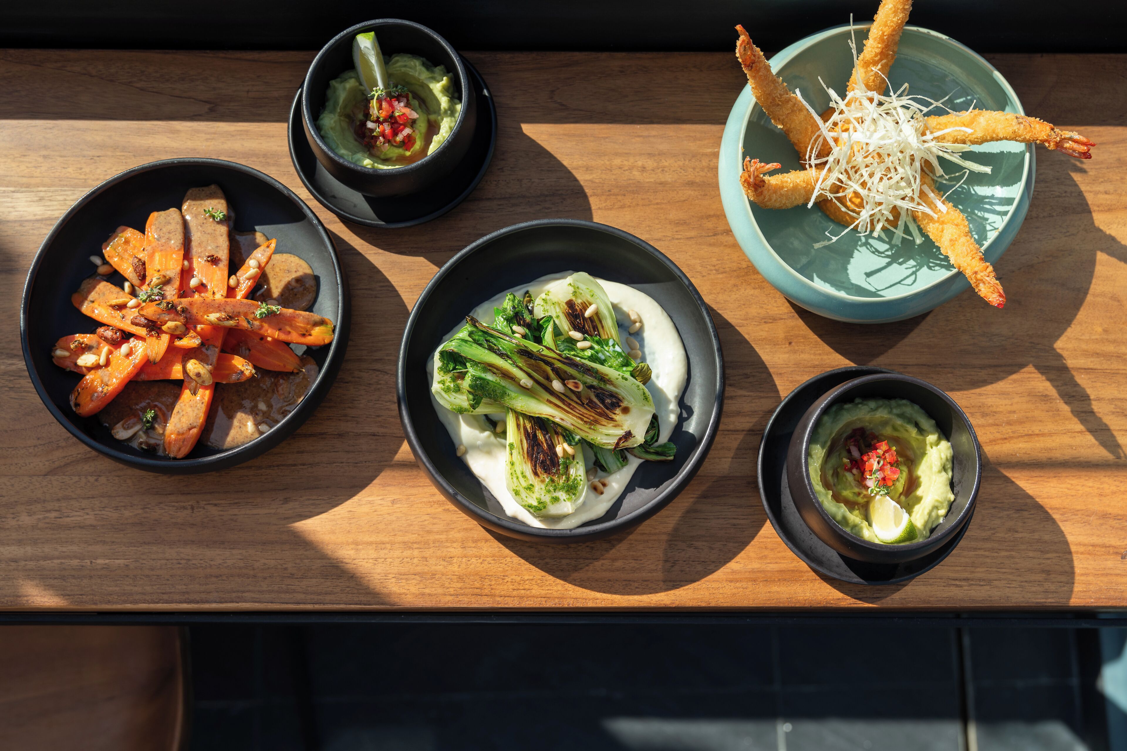 A wooden table with four bowls of food, including roasted carrots, a green vegetable dish, and a bowl with a creamy substance topped with a red garnish.