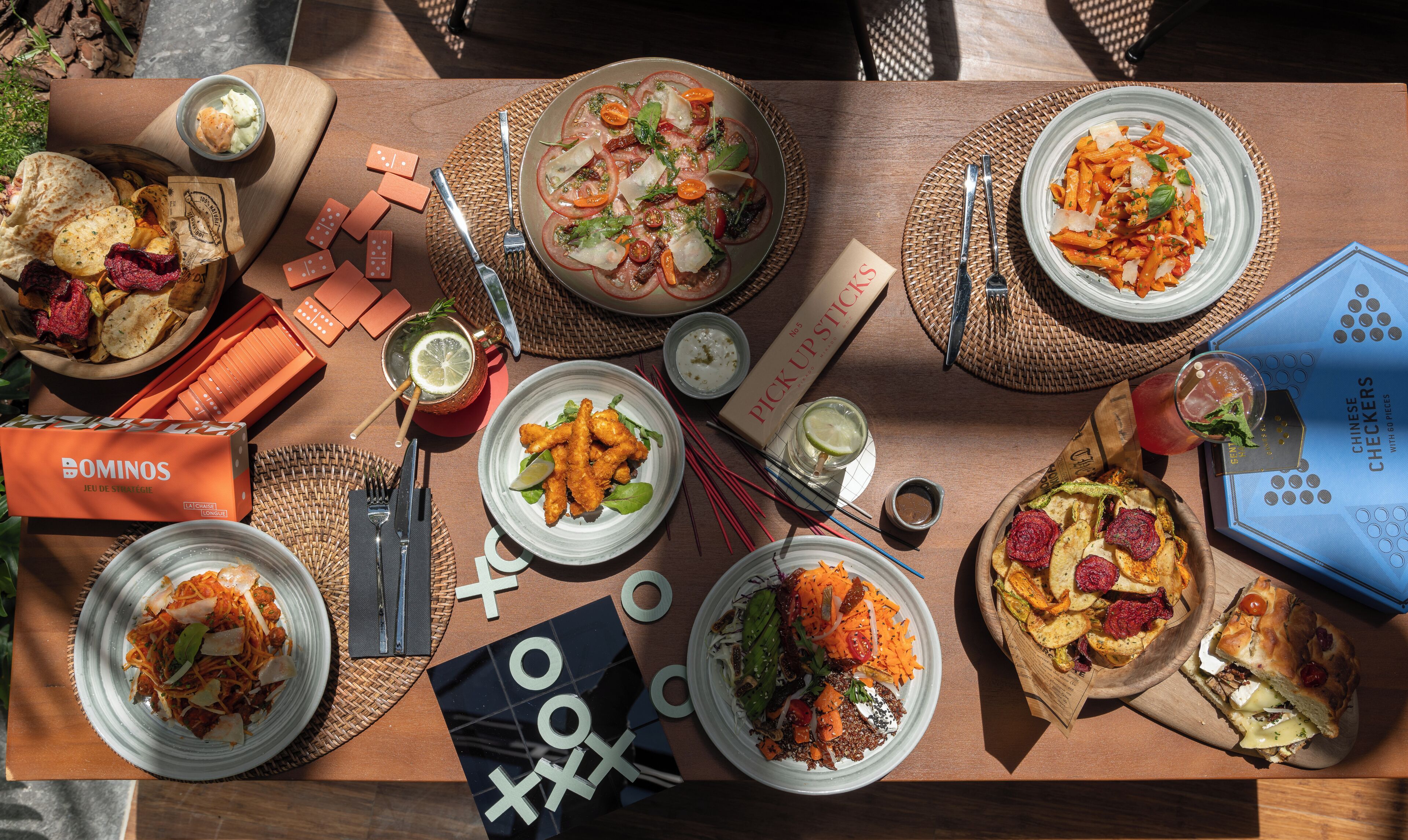Una mesa servida con diversos platos, incluyendo pasta, ensaladas y pan, además de salsas y una caja de juegos.