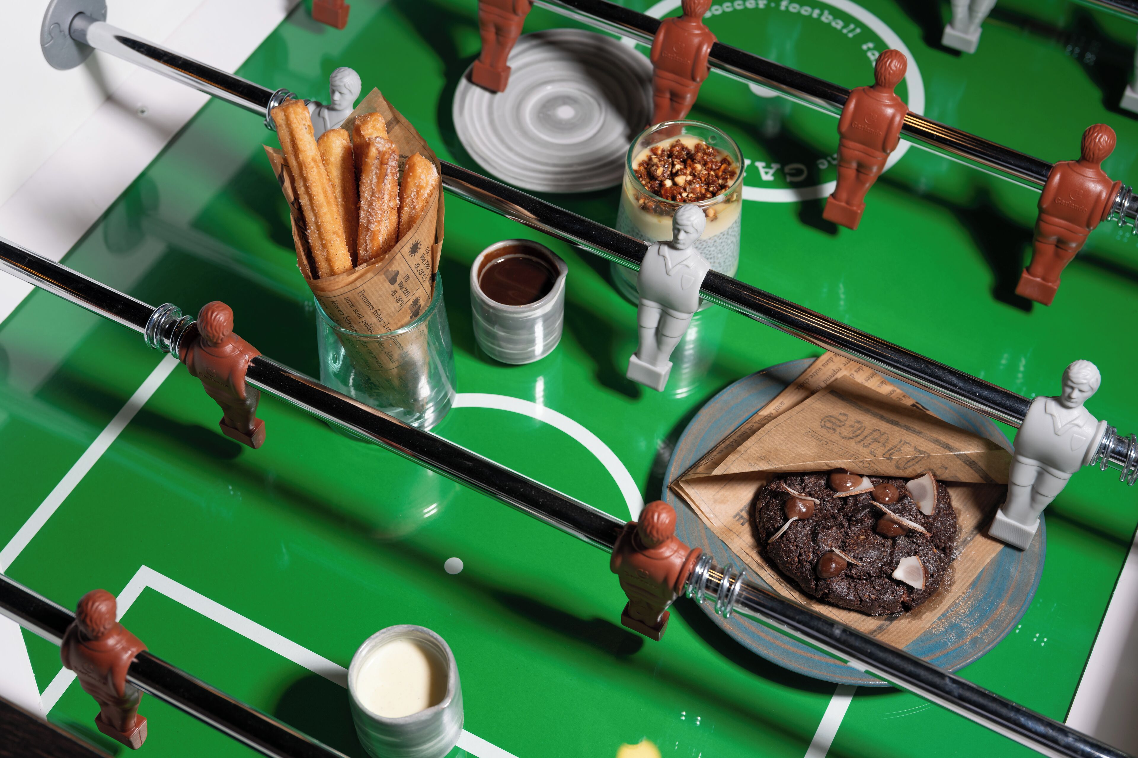 A tabletop with a green surface, a small wooden board with a chocolate dessert, a glass with a creamy beverage, and a container of what appears to be a chocolate spread.